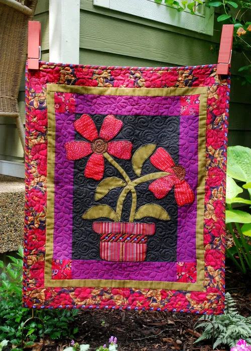Mini Quilt Patterns You'll Want to Make This Weekend 10 A colorful mini quilt with a floral design features two red flowers with green stems and leaves in a striped pot, bordered by purple and red patterned fabric, hanging outdoors on a clothesline with clothespins. Perfect inspiration for mini quilt patterns!.
