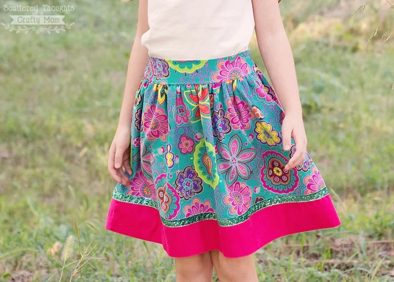 15 Easy Elastic Waist Skirt Patterns You Can Sew Today (All Free) 16 A child wearing a colorful floral skirt with a bright pink hem stands outdoors on grass. The skirt features an elastic waist, perfect for easy skirt patterns inspired by free skirt patterns. The child's upper body is out of frame.