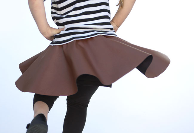 15 Easy Elastic Waist Skirt Patterns You Can Sew Today (All Free) 17 A child wearing a striped shirt and brown skirt twirls, causing the skirt to flare out—a perfect showcase for easy skirt patterns. The background is plain white.