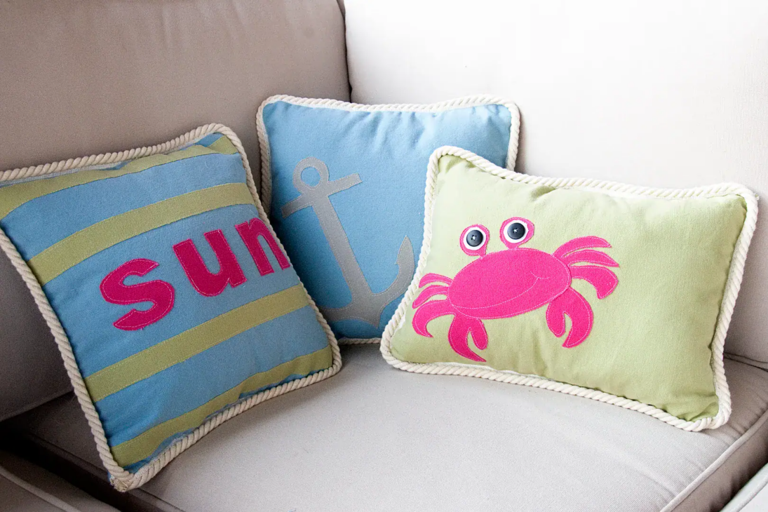 Refresh Your Home with These Summer Pillow Sewing Ideas (free patterns) 7 Three decorative pillows on a white couch; one with “Sun” text, one with an anchor design, and one with a pink crab illustration—perfect for a summer pillow sewing project or an easy home refresh.