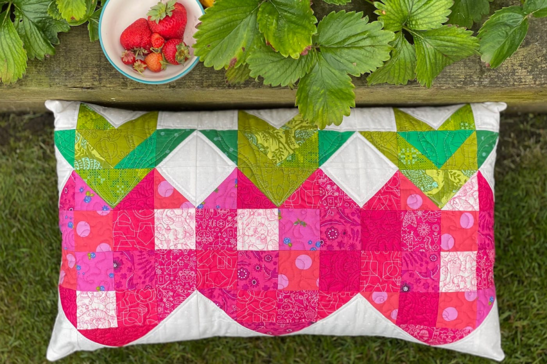 Refresh Your Home with These Summer Pillow Sewing Ideas (free patterns) 11 A quilted pillow with a pink and green strawberry pattern—perfect for a summer pillow sewing project—lies on grass near a wooden table holding a bowl of strawberries and fresh strawberry plant leaves, offering an inviting home refresh.