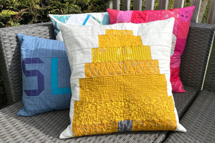 Refresh Your Home with These Summer Pillow Sewing Ideas (free patterns) 6 Three decorative pillows on a wicker bench provide the perfect inspiration for your next summer pillow sewing project. The front pillow features a cheerful yellow geometric quilt pattern, while the two behind display charming patchwork designs in blue and pink tones—ideal choices for a home refresh. In the background, a wooden railing and lush greenery complete this inviting scene. Discover free patterns to recreate these looks in your own space.