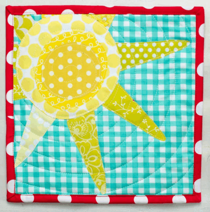 Mini Quilt Patterns You'll Want to Make This Weekend 11 A quilted fabric square features a yellow sun with patterned rays on a blue and white gingham background. The red border with white polka dots highlights charming mini quilt patterns, and circular stitching adds texture.