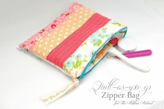 A colorful, quilted zipper bag with floral and striped bag patterns is unzipped, showing scissors and sewing tools inside. The bag rests on a white surface with "Quilt-As-You-Go Zipper Bag" text beside it.