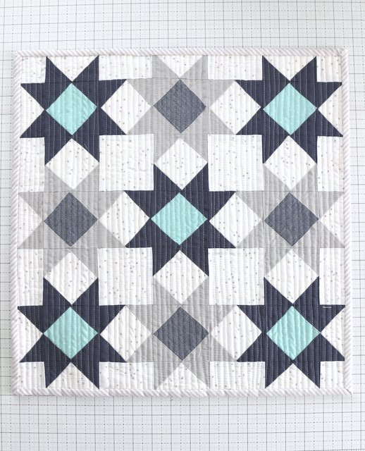 Mini Quilt Patterns You'll Want to Make This Weekend 12 A quilt with a geometric pattern featuring navy blue, light blue, gray, and white fabrics arranged in star and diamond shapes on a grid background—perfect inspiration for mini quilts or unique quilt patterns.
