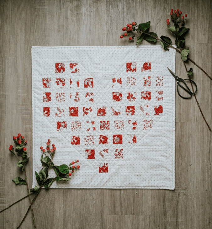 Mini Quilt Patterns You'll Want to Make This Weekend 13 A quilt with a heart pattern made of small red and white fabric squares is laid on a wooden floor, surrounded by branches with red berries and green leaves, showcasing charming mini quilt patterns, with a pair of scissors on the right side.