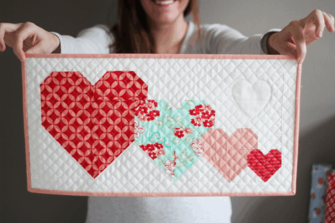 Mini Quilt Patterns You'll Want to Make This Weekend 14 A person holds a quilted rectangular panel featuring four patchwork hearts in red, pink, and blue quilt patterns on a white background with a pale pink border. The person is smiling but their face is out of focus—perfect inspiration for mini quilts.