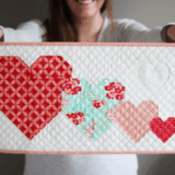 A person holds a quilted rectangular panel featuring four patchwork hearts in red, pink, and blue quilt patterns on a white background with a pale pink border. The person is smiling but their face is out of focus—perfect inspiration for mini quilts.