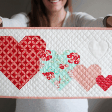 A person holds a quilted rectangular panel featuring four patchwork hearts in red, pink, and blue quilt patterns on a white background with a pale pink border. The person is smiling but their face is out of focus—perfect inspiration for mini quilts.