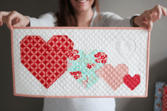 A person holds a quilted rectangular panel featuring four patchwork hearts in red, pink, and blue quilt patterns on a white background with a pale pink border. The person is smiling but their face is out of focus&mdash;perfect inspiration for mini quilts.