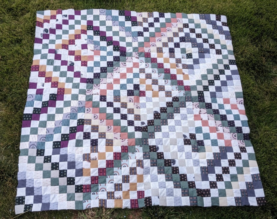 Free Online Creative Summit for Exploring New Hobbies 4 A colorful patchwork quilt with a geometric pattern of small squares in various shades of purple, green, blue, pink, and white is spread out on green grass—perfect for relaxing outdoors while Exploring New Hobbies or attending a Free Creative Summit online.