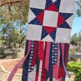 A quilted patriotic Seasonal Ribbon Banner with a red, white, and blue star hangs from a tree, with ribbon-like fabric strips below. This vibrant seasonal decoration adds festive charm to the desert landscaping and trees in the background—perfect to celebrate every season.