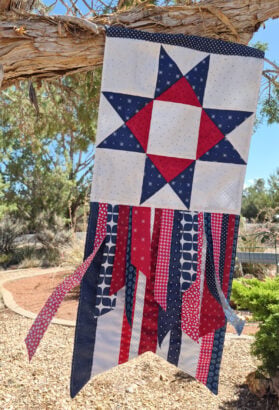 A quilted patriotic Seasonal Ribbon Banner with a red, white, and blue star hangs from a tree, with ribbon-like fabric strips below. This vibrant seasonal decoration adds festive charm to the desert landscaping and trees in the background—perfect to celebrate every season.