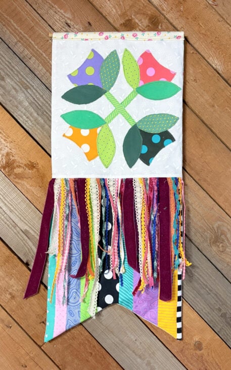 Seasonal Ribbon Banner – A Fun Way to Celebrate Every Season 1 A quilted wall hanging with four colorful flower shapes, green leaves, and stems brightens any room. Below, a vibrant Seasonal Ribbon Banner—with multicolored fabric strips and ribbons—hangs over a patchwork base on a wood plank floor, creating the perfect seasonal decoration to celebrate every season in style.
