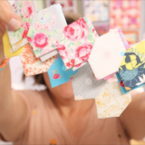 A person holds up several small, colorful, patterned fabric squares—perfect for English Paper Piecing—displaying them closely to the camera. The focus is on the fabrics, which appear to be cut using an AccuQuilt Honeycomb Die, while the person in the background is blurred.