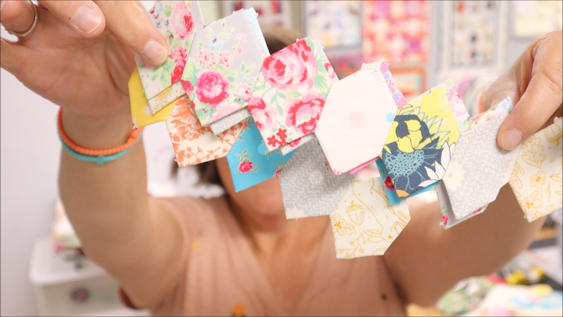English Paper Piecing Honeycomb-1" Finished Sides Die AccuQuilt 1 A person holds up several small, colorful, patterned fabric squares—perfect for English Paper Piecing—displaying them closely to the camera. The focus is on the fabrics, which appear to be cut using an AccuQuilt Honeycomb Die, while the person in the background is blurred.