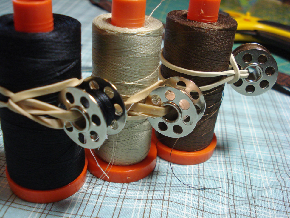 Bobbin Storage Ideas That Will Finally Keep Those Threads Under Control 2 Three spools of thread—black, beige, and brown—each with a metal bobbin tied on using a rubber band, are placed on a blue plaid fabric background, offering a simple yet effective bobbin storage solution for your sewing room organization.