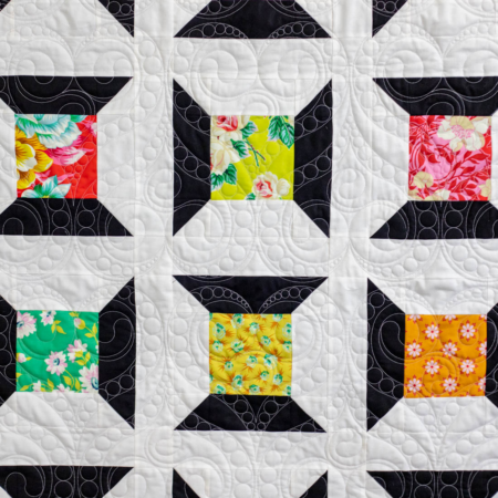 A quilt with a geometric pattern featuring black, white, and various colorful floral squares, inspired by free charm pack quilt patterns. The stitching creates swirling designs across the fabric.