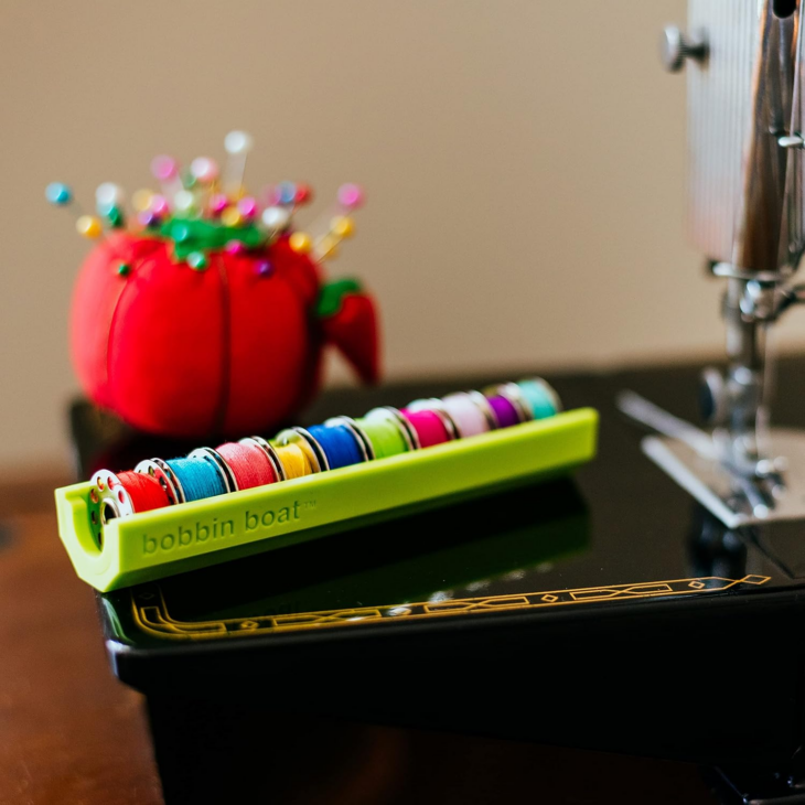 Bobbin Storage Ideas That Will Finally Keep Those Threads Under Control 5 A green “bobbin boat” designed for bobbin storage holds colorful sewing machine bobbins next to a red tomato pincushion with pins and a sewing machine on a dark table, offering clever thread organization inspiration for your sewing room ideas.