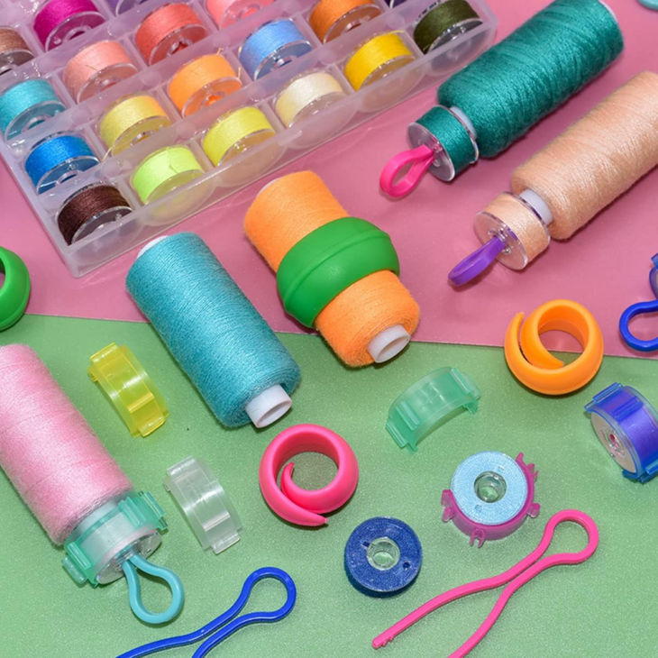 Bobbin Storage Ideas That Will Finally Keep Those Threads Under Control 7 Colorful sewing threads, bobbins, and plastic sewing tools are arranged on a pink and green background, showcasing creative thread organization. There is also a clear plastic case with multiple bobbins filled with different colored threads, serving as an excellent bobbin organizer.