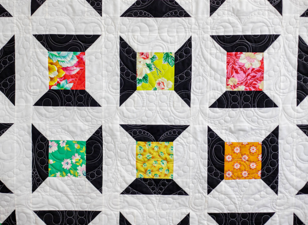 A quilt with a geometric pattern featuring black, white, and various colorful floral squares, inspired by free charm pack quilt patterns. The stitching creates swirling designs across the fabric.