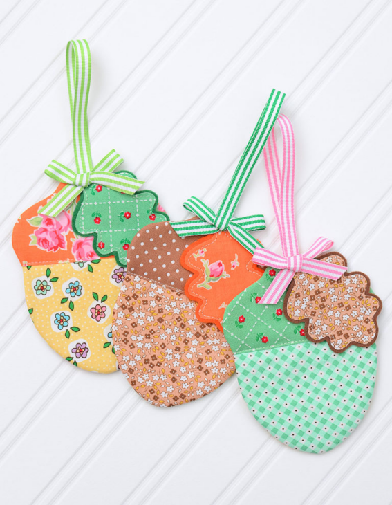 Fall Appliqué Patterns That Are Perfect for Your Fabric Scraps 11 Three fabric acorn ornaments with floral, polka dot, and gingham patterns, each featuring fall applique details and a colorful ribbon loop for hanging, displayed on a white background.
