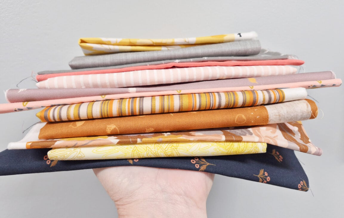 Switchback Mystery Quilt Along – Get Ready to Sew! 3 A hand holds a stack of folded fabric squares in yellow, gray, pink, and dark blue for a Switchback Mystery Quilt sewing project.