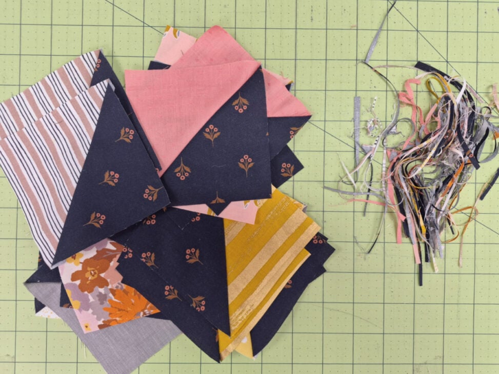 Switchback Mystery Quilt Along – Get Ready to Sew! 3 A stack of colorful fabric triangles lies on a green cutting mat, next to a pile of thin, multicolored fabric scraps and trimmings—perfect for anyone eager to sew along with the Switchback Mystery Quilt or join a vibrant Quilt Along.