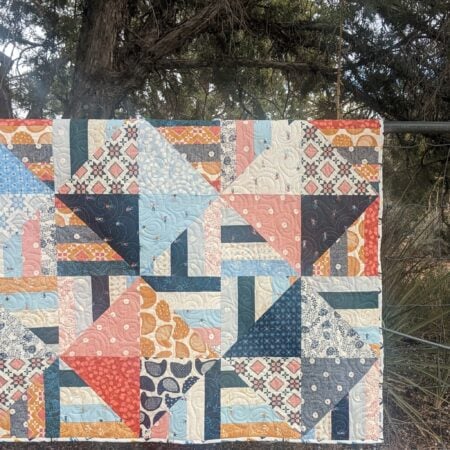A geometric Jelly Roll quilt featuring squares and triangles in blue, peach, orange, and white patterns, hung on a metal fence.