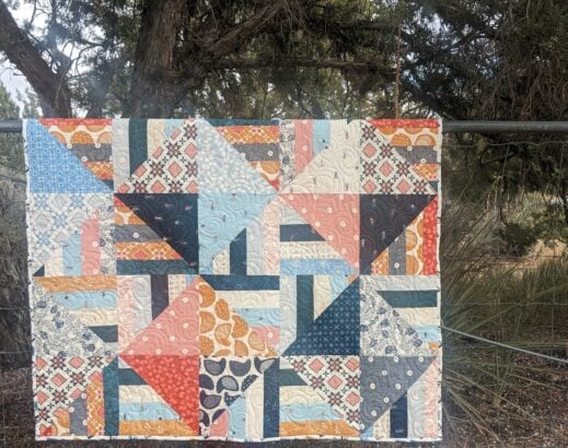 A geometric Jelly Roll quilt featuring squares and triangles in blue, peach, orange, and white patterns, hung on a metal fence.