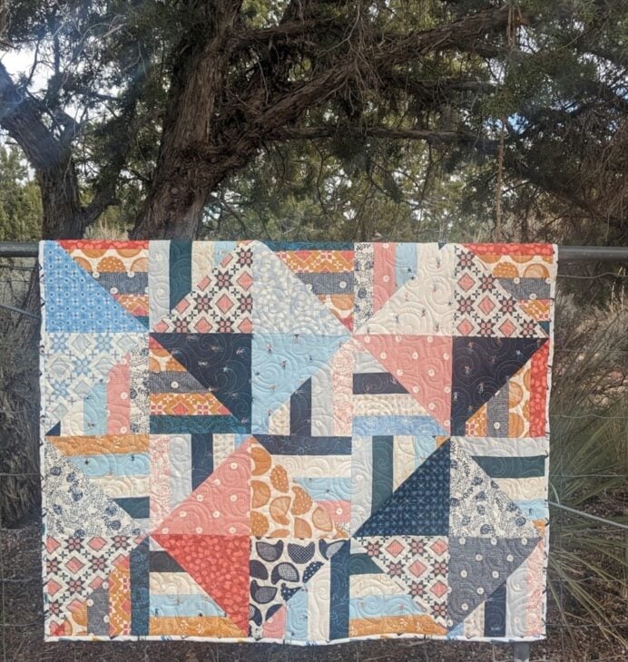 Jelly Roll + Layer Cake Quilt – Mix, Match & Sew It Up 7 A Jelly Roll quilt featuring a geometric pattern of triangles and rectangles in various blue, pink, orange, and cream patterned fabrics, hanging on a wire fence in front of a tree.