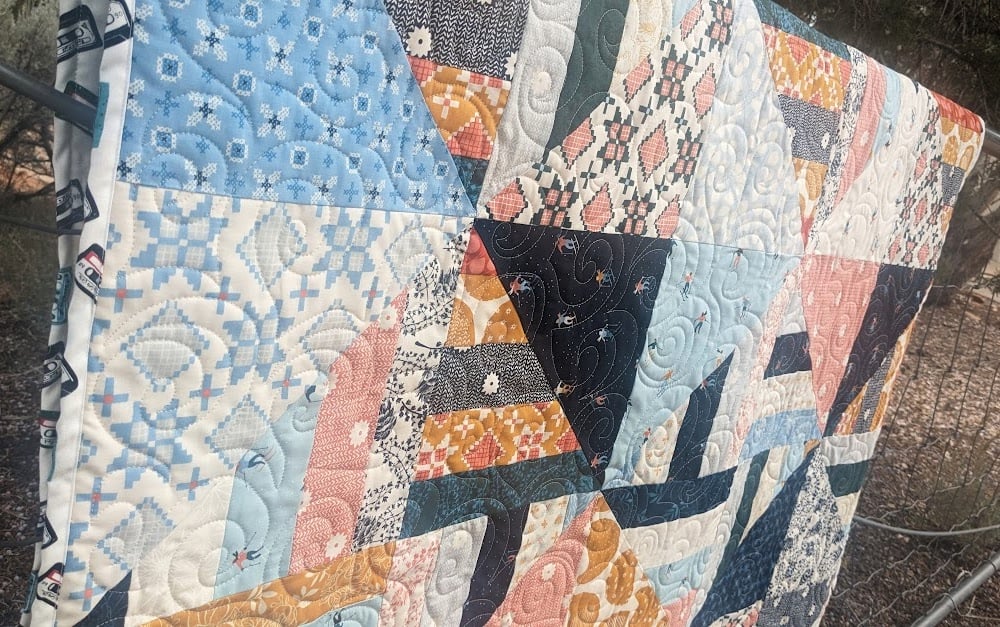 Jelly Roll + Layer Cake Quilt – Mix, Match & Sew It Up 5 A Jelly Roll quilt made of various blue, orange, and black patterned fabric triangles, featuring swirling machine-stitched quilting patterns, draped over a metal fence.