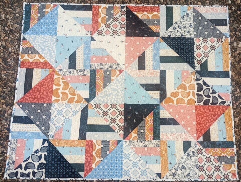 Jelly Roll + Layer Cake Quilt – Mix, Match & Sew It Up 4 A Layer Cake quilt featuring a geometric design of triangles and strips in a variety of patterned fabrics in blue, red, orange, and cream.