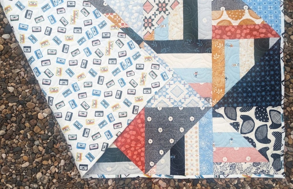 Jelly Roll + Layer Cake Quilt – Mix, Match & Sew It Up 3 A Jelly Roll quilt with a geometric patchwork design on one side and a repeating cassette tape pattern on the other, lying on a bed of pebbles.