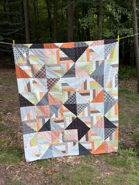 Jelly Roll + Layer Cake Quilt – Mix, Match & Sew It Up 1 A colorful patchwork Jelly Roll quilt with orange, black, white, and gray patterns hangs on a line outdoors, framed by a forested background and leafy ground.