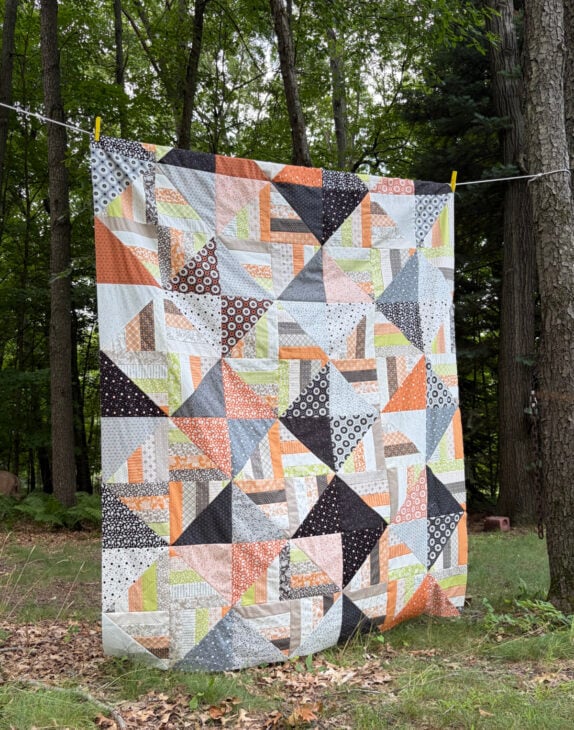 Jelly Roll + Layer Cake Quilt – Mix, Match & Sew It Up 2 A Layer Cake patchwork quilt with a geometric pattern of triangles and rectangles in orange, gray, white, and black, hanging on a clothesline between two trees.