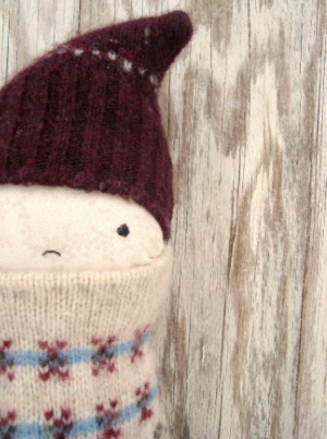 A handmade soft doll with a small, neutral face, wearing a maroon knitted hat and a cream sweater with blue and red patterns, is positioned against a rustic wooden background&mdash;start here for charming, handcrafted decor.